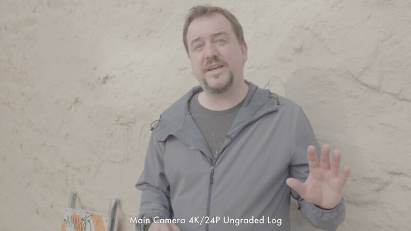 A man in a gray jacket stands outdoors in front of a beige textured wall, holding one hand up mid-gesture. White text at the bottom reads, “Main Camera 4K/24P Ungraded Log.”.