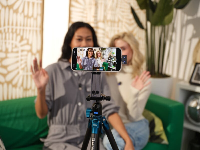 Two women sit on a green couch waving at a smartphone camera on a tripod, filming themselves. The phone screen displays their image as they record a video in a brightly lit, modern room with plants and decorative panels.