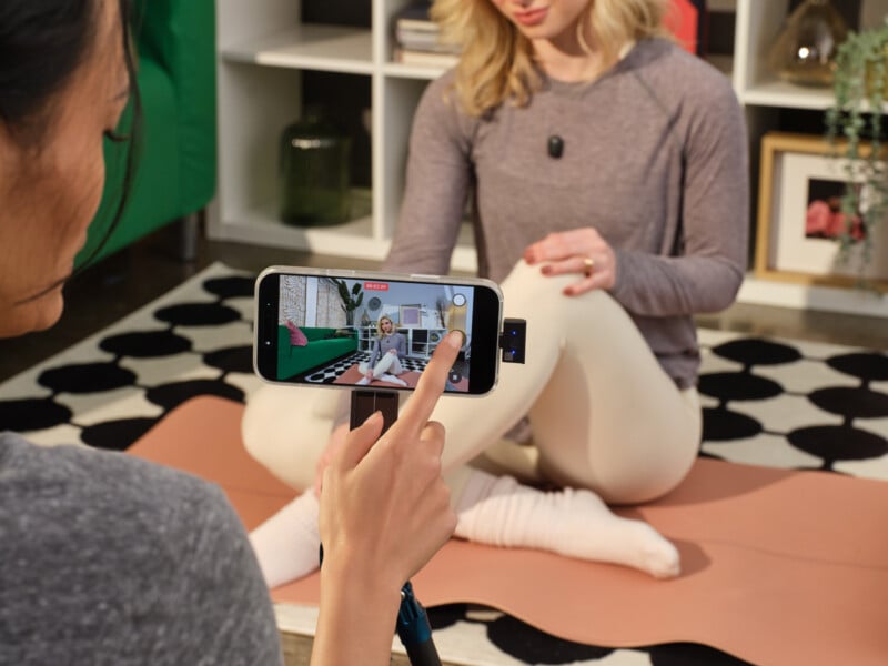A woman records another woman sitting on a yoga mat using a smartphone on a tripod. The phone screen shows the seated woman as she demonstrates a stretch in a cozy indoor space.