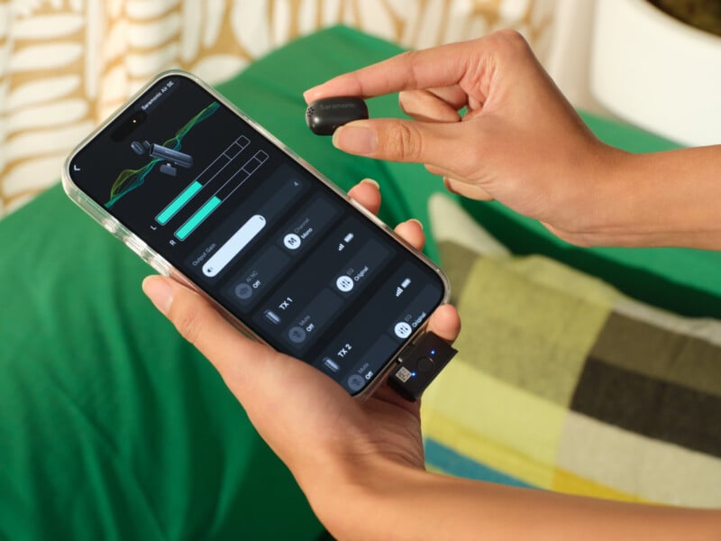 A person holds a wireless earbud in one hand and a smartphone in the other, displaying an audio control app on the screen. A green blanket and plaid pillow are visible in the background.