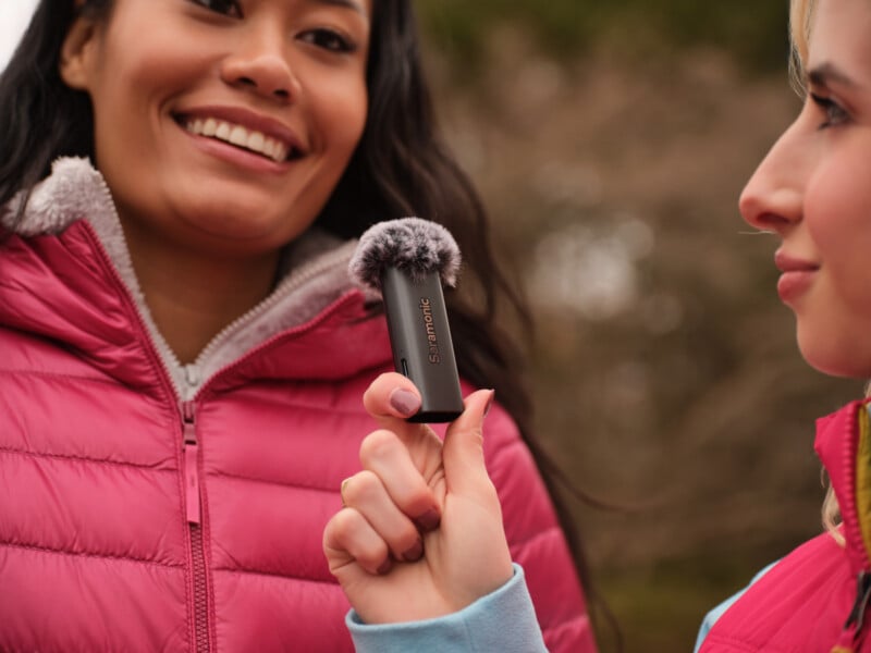 Two women outdoors, one smiling and wearing a pink jacket, while the other holds a small black Saramonic microphone with a furry windscreen close to her.