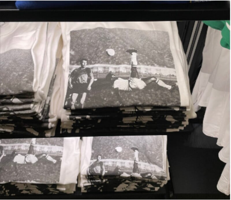 Stacks of folded white t-shirts on a shelf, featuring a black-and-white print of a soccer player performing a bicycle kick with another player and a soccer ball visible in the background.