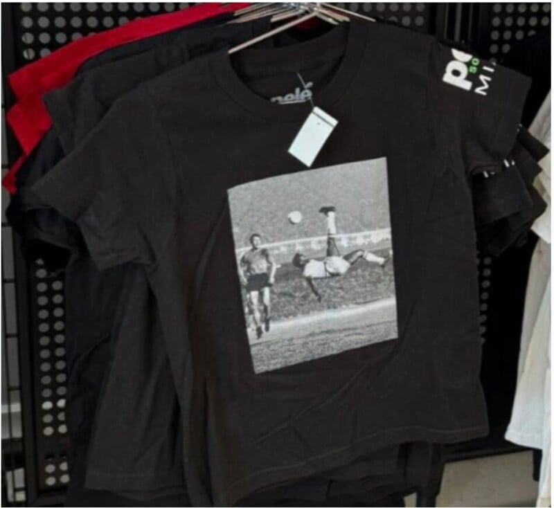 A black t-shirt hangs on a rack, printed with a vintage black-and-white image of a soccer player performing a bicycle kick as another player watches. Other t-shirts in various colors are visible in the background.