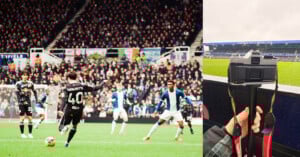 A soccer player in a black jersey prepares to kick the ball during a match, while defenders in blue and white watch. Beside the field, a hand holds a camera on a monopod, with the stadium and crowd visible.