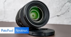 A close-up of a camera lens placed on a table with its cap underneath, in front of a light grey textured background. "PetaPixel Reviews" text is displayed in the lower left corner.