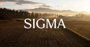 A cultivated field at sunrise with misty trees and mountains in the background. Large white text reading "SIGMA" is centered over the landscape.