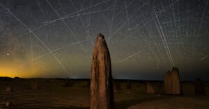 Tall limestone pillars rise from sandy desert terrain under a clear night sky filled with stars, intersected by bright streaks of light from moving satellites or meteors, creating a web-like effect.