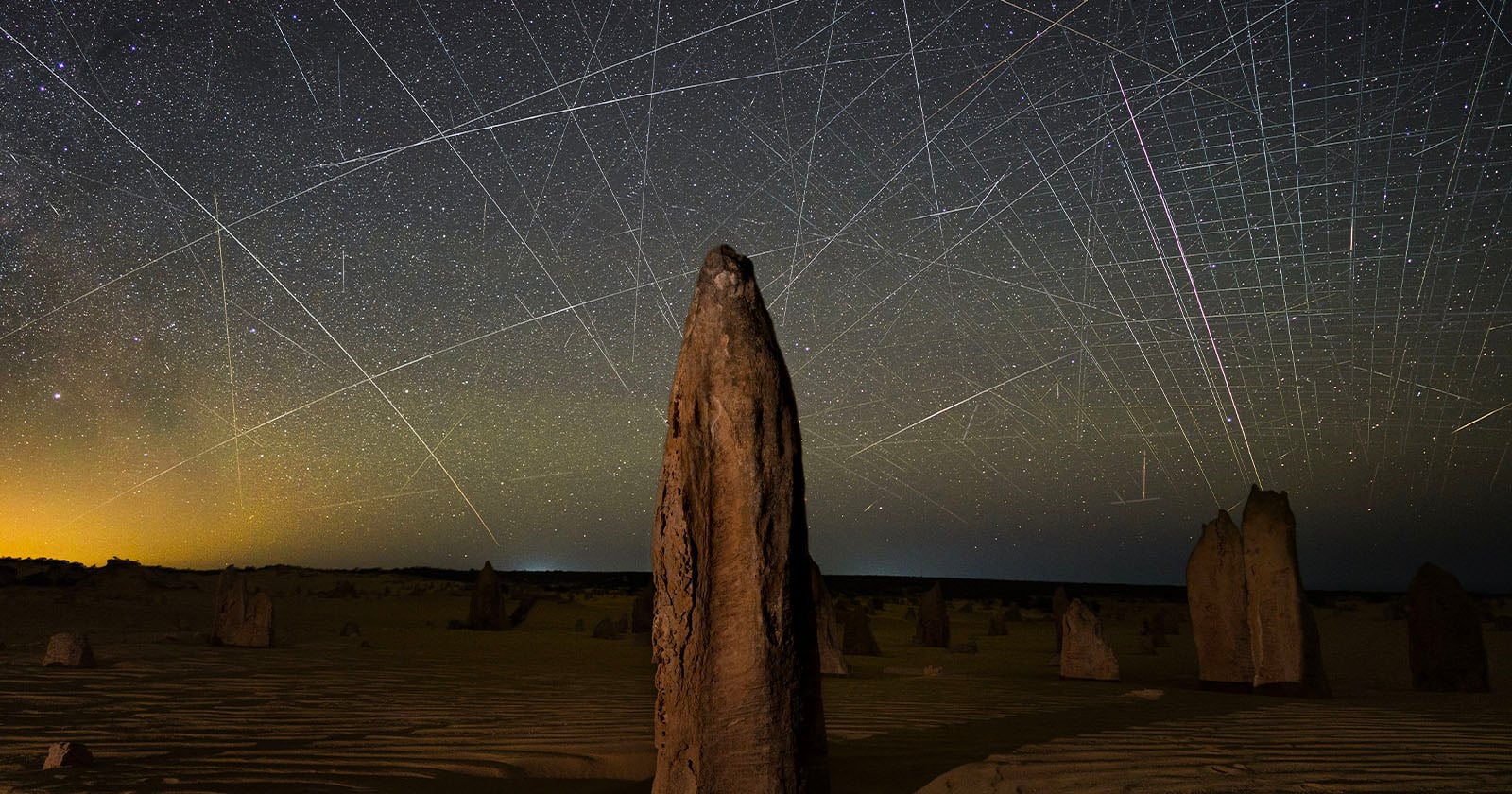 Tall limestone pillars rise from sandy desert terrain under a clear night sky filled with stars, intersected by bright streaks of light from moving satellites or meteors, creating a web-like effect.