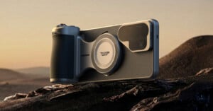 A smartphone with a large camera and ergonomic grip attachment rests on a rocky surface with a desert landscape in the background during sunset.