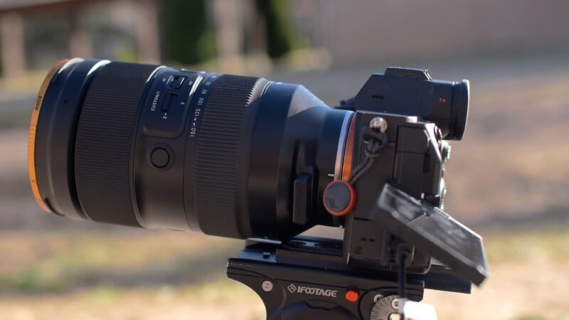 Primer plano de una cámara profesional con su gran teleobjetivo montado en un cabezal de trípode iFootage, colocado al aire libre con un fondo natural borroso.