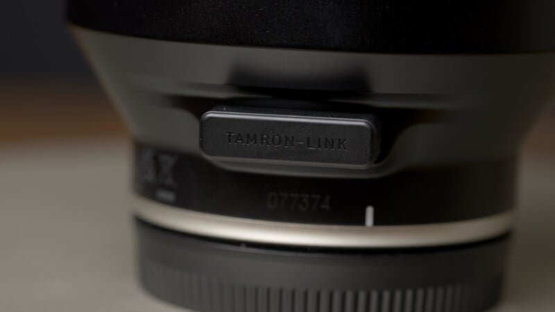 Primer plano de la lente de la cámara con etiqueta de cubierta de plástico negro "ENLACE Tamron," Mostrando parte de la montura del objetivo y el número de serie 077374 debajo.