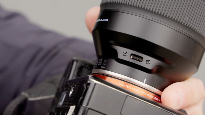 Primer plano de las manos de una persona sujetando la lente de una cámara al cuerpo de una cámara digital, mostrando el puerto USB en la lente y el texto 