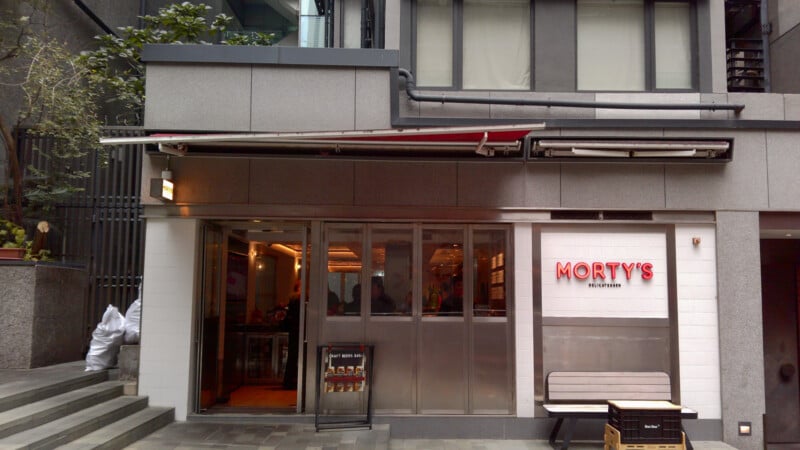 Street view of Morty's Delicatessen, featuring a modern gray and white exterior, glass doors, a small bench, and a menu board beside the entrance. The restaurant’s name is prominently displayed in red letters.
