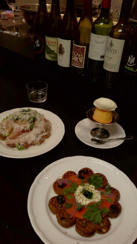 A table set with two plates of food, a dessert with ice cream, a glass of water, and several bottles of wine in the background. The dishes are artfully arranged and garnished with herbs and cheese.