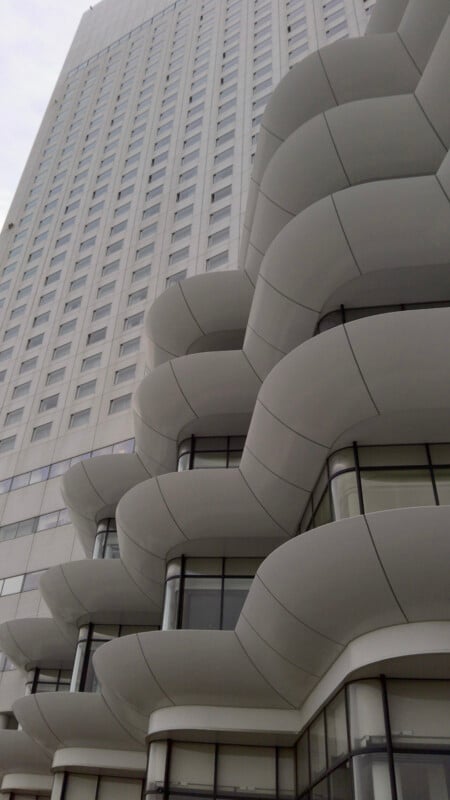 A modern high-rise building with curved, layered white balconies and large windows, viewed from below against a cloudy sky.