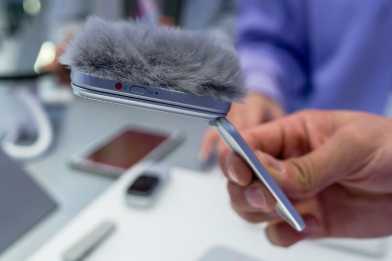 Una persona sostiene un teléfono inteligente plegable con un parabrisas gris esponjoso en la parte superior, posiblemente para grabar audio. El fondo está borroso, mostrando otros dispositivos electrónicos sobre la mesa.