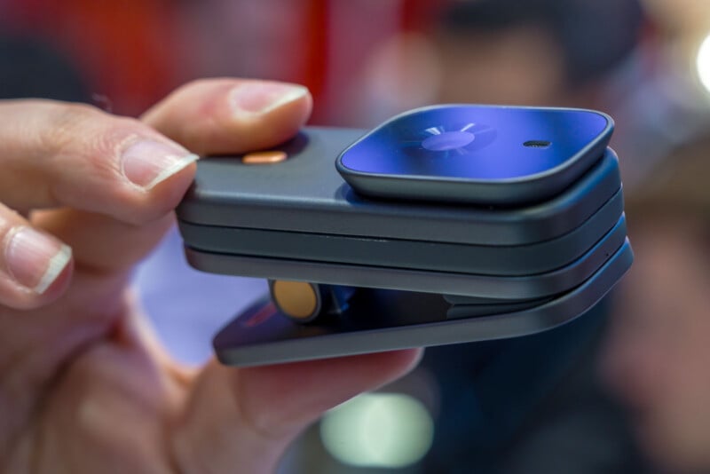 Primer plano de una mano que sostiene un dispositivo pequeño y compacto con una lente o sensor redondo y elegante en la parte superior y un cuerpo rectangular en capas con botones a los lados. El fondo está borroso.