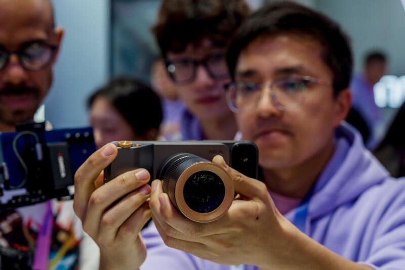Un hombre con una sudadera con capucha morada sostiene un teléfono inteligente con una gran lente de cámara y enfoca atentamente, mientras otros se paran cerca y observan más de cerca el ajetreado ambiente interior.