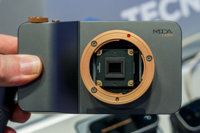 Un primer plano de una mano que muestra un módulo de cámara con lente MODA con un sensor de imagen de luz visible y una montura de lente dorada en un evento tecnológico.
