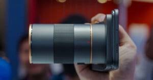 A hand holding a smartphone with a large professional camera lens attached, showing a close-up side view. The background is blurred with indistinct people and colorful lights.