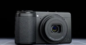 A black compact digital camera with a textured grip and a protruding lens, labeled "GR LENS f=18.3mm 1:2.8," is placed on a flat surface against a dark background.