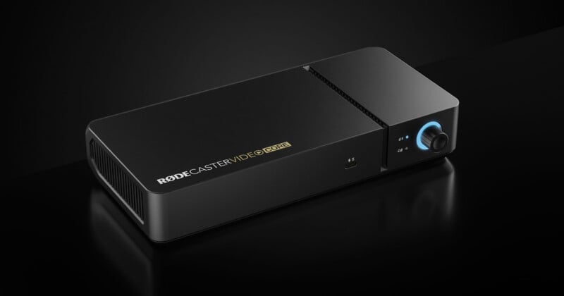 A black rectangular audio device labeled "RODECASTER VIDEO CORE" with a blue-lit knob, buttons, and vents, set against a dark background.