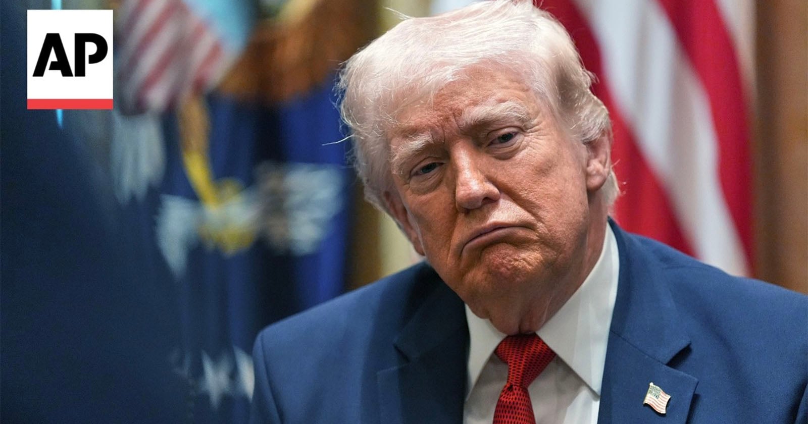 A man in a suit with an American flag pin sits with a serious expression. Behind him are blurred flags, including the U.S. flag. The Associated Press “AP” logo is in the upper left corner.