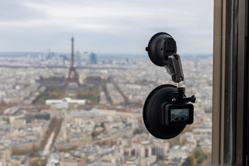 A small camera with suction mounts is attached to a window, filming a cityscape with the Eiffel Tower in the background on a cloudy day.