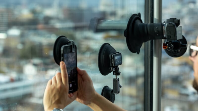 A person uses a smartphone mounted on a suction cup to photograph a cityscape through a window. Nearby, an action camera and a DSLR camera are also attached to the glass with suction mounts.