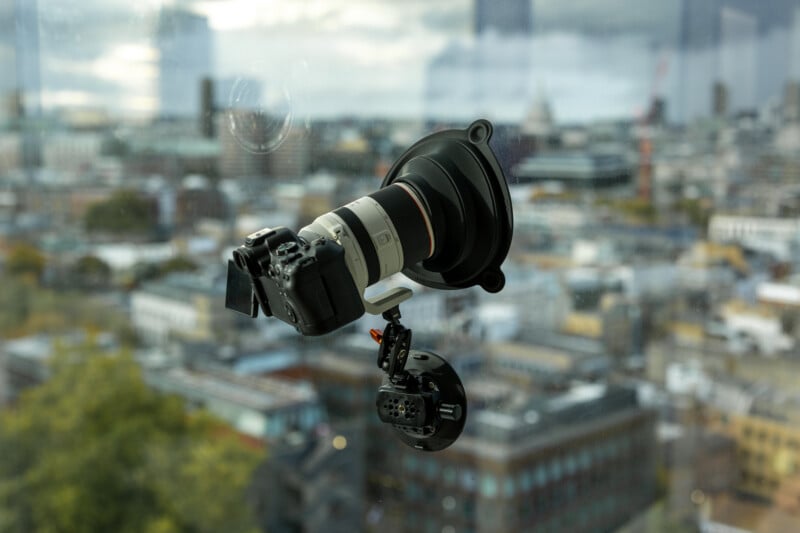 A camera with a telephoto lens is mounted to a window using a suction cup mount, overlooking a blurred cityscape in the background.