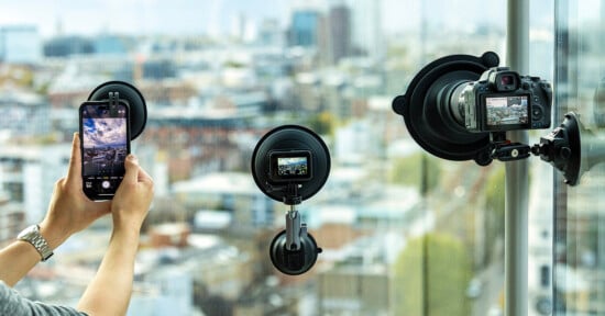 A person takes a photo of a cityscape through a window using a smartphone, while two cameras are mounted on the glass with large suction cup mounts, also filming or photographing the view.