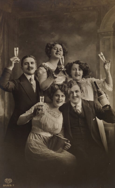Five people in vintage clothing smile and raise glasses of champagne in a celebratory toast. The image is sepia-toned, giving it an old-fashioned, early 20th-century feel. The group appears joyful and festive.