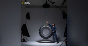 A man in a studio setting stands next to a large tire hanging from the ceiling by a rope. He is touching the tire, and professional lighting equipment is visible around him. The National Geographic logo appears in the top left corner.