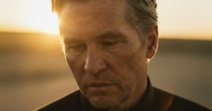 A middle-aged man with gray hair looks down with a somber expression, standing outdoors at sunset with warm sunlight illuminating his face.