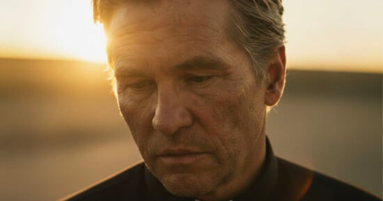 A middle-aged man with gray hair looks down with a somber expression, standing outdoors at sunset with warm sunlight illuminating his face.