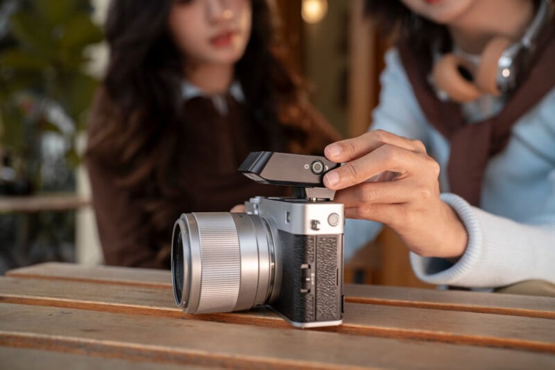 A person’s hand adjusts the viewfinder of a silver digital camera on a wooden table, with two people sitting in the background, slightly out of focus.