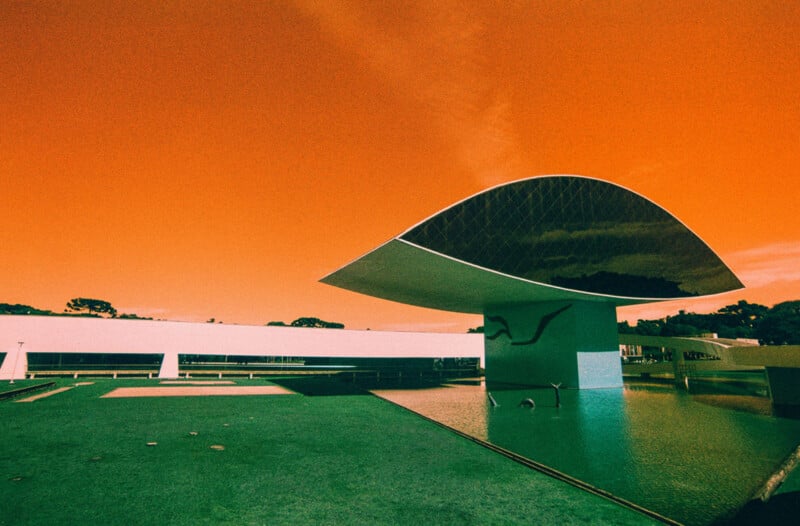 A modern building with a curved, reflective roof stands over a shallow pool under an intense orange sky, creating a surreal atmosphere. The structure features geometric lines and a minimalist design.