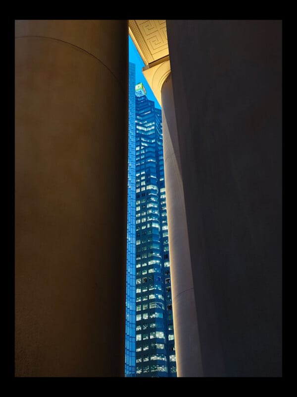 A modern glass skyscraper is seen through a narrow gap between two large, classical columns at dusk, with the building’s windows illuminated against a deep blue sky.