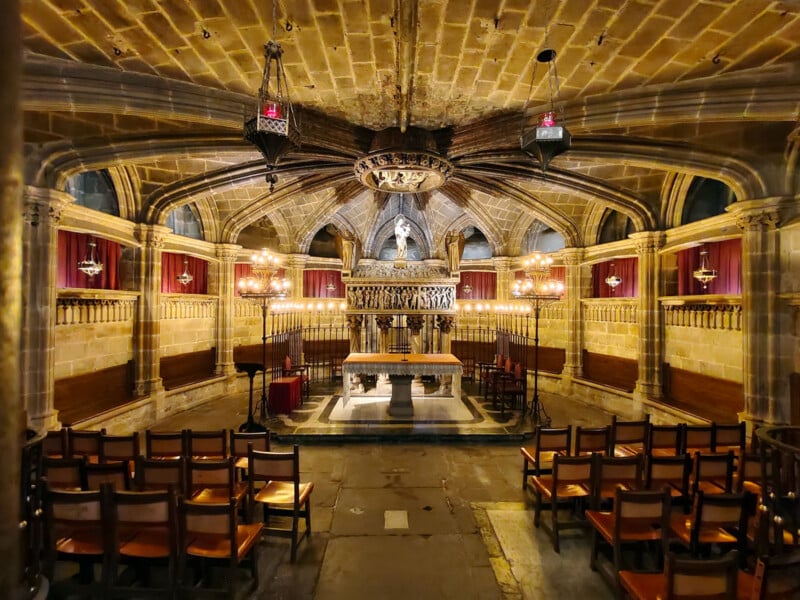 A stone chapel with vaulted ceilings, ornate chandeliers, wooden chairs, a central altar, red drapes along the walls, and a statue above the altar, all warmly lit and richly decorated.