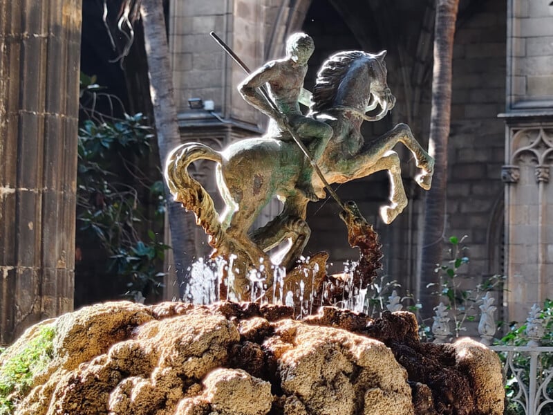A bronze statue of a rider on a rearing horse holding a spear, mounted on a rocky fountain with water spraying upward, set against a historic stone building and lush greenery.