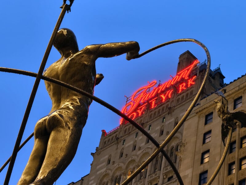 Bronze statue of a figure holding a rope in front of the Fairmont Royal York hotel, with its red neon sign glowing against a clear blue sky.