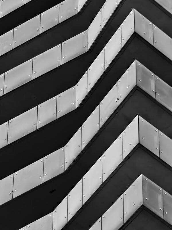 Black and white photo showing a close-up of zigzagging, geometric balconies on a modern building, with strong lines and shadows creating an abstract, repetitive pattern.