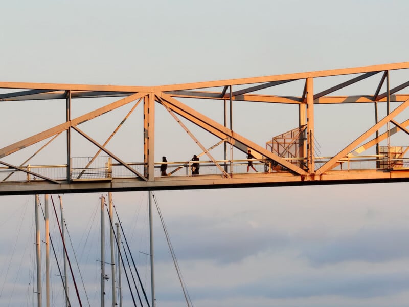 People walking across an elevated pedestrian bridge at sunset, with the structure illuminated by warm light. Below, the tops of sailboat masts are visible against a cloudy sky.