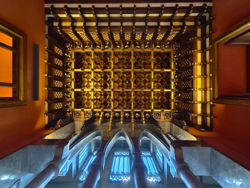 Ornate ceiling with intricate golden latticework, viewed from below, surrounded by red and orange walls, arched windows, and blue-toned marble columns.
