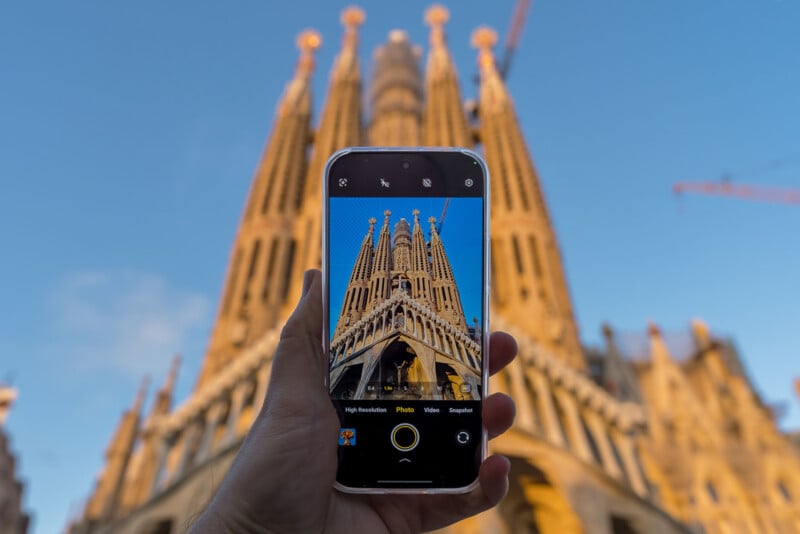 A hand holds a smartphone, capturing a photo of a tall, ornate cathedral with spires, possibly the Sagrada Familia, against a blue sky. The phone screen shows the detailed architecture in focus.