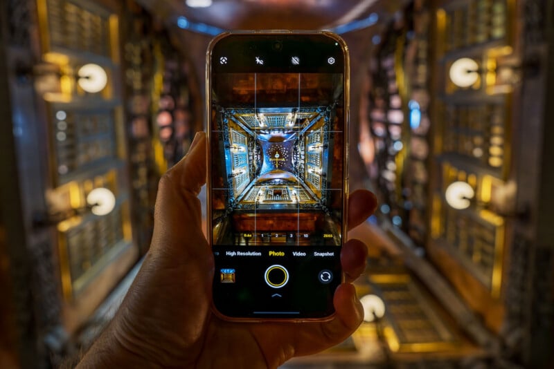 A hand holds a smartphone, capturing a photo of an ornate, symmetrical ceiling with warm lighting and intricate patterns; the phone screen mirrors the detailed architectural view above.