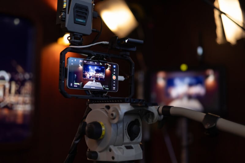 A camera rig with a smartphone attached is filming a studio set with bright lights and blurred background screens, capturing a production scene.