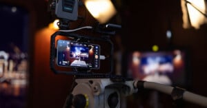 A professional video camera rig with a smartphone mounted on a tripod captures a studio scene with bright lights and blurred background. The phone’s screen displays the live camera view.