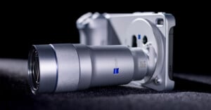 A close-up view of a modern digital camera with a large silver lens, lying on a dark textured surface with soft lighting highlighting its sleek metallic design.