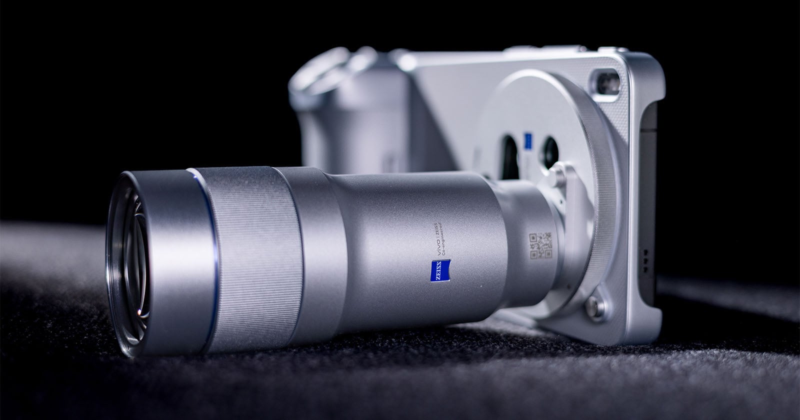 A close-up view of a modern digital camera with a large silver lens, lying on a dark textured surface with soft lighting highlighting its sleek metallic design.